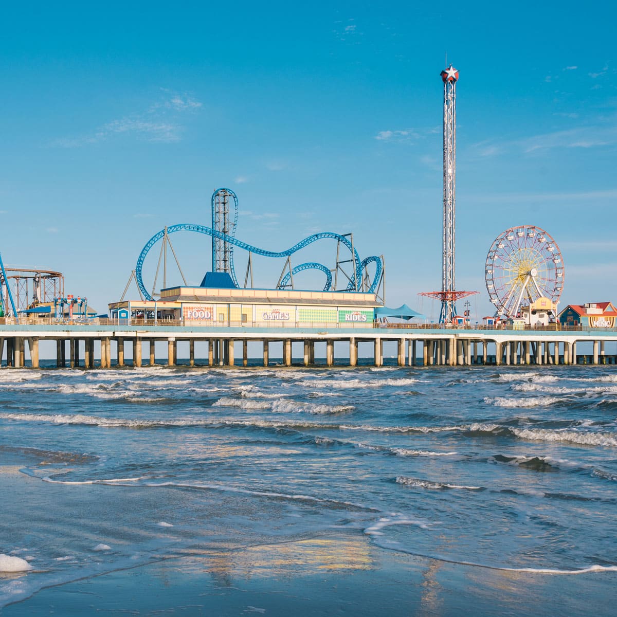A seaside amusement park on a pier features a blue roller coaster, tall drop tower, and Ferris wheel above the ocean waves on a clear, sunny day - setting the stage for Ironman 70.3 Texas.